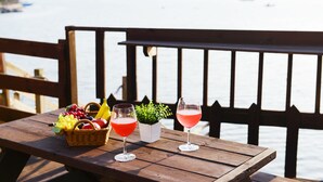 Ein Holztisch mit einem Korb voller Früchte, zwei Gläser rosa Getränke und einem kleinen Blumentopf auf einem Balkon mit Blick auf ein Gewässer.