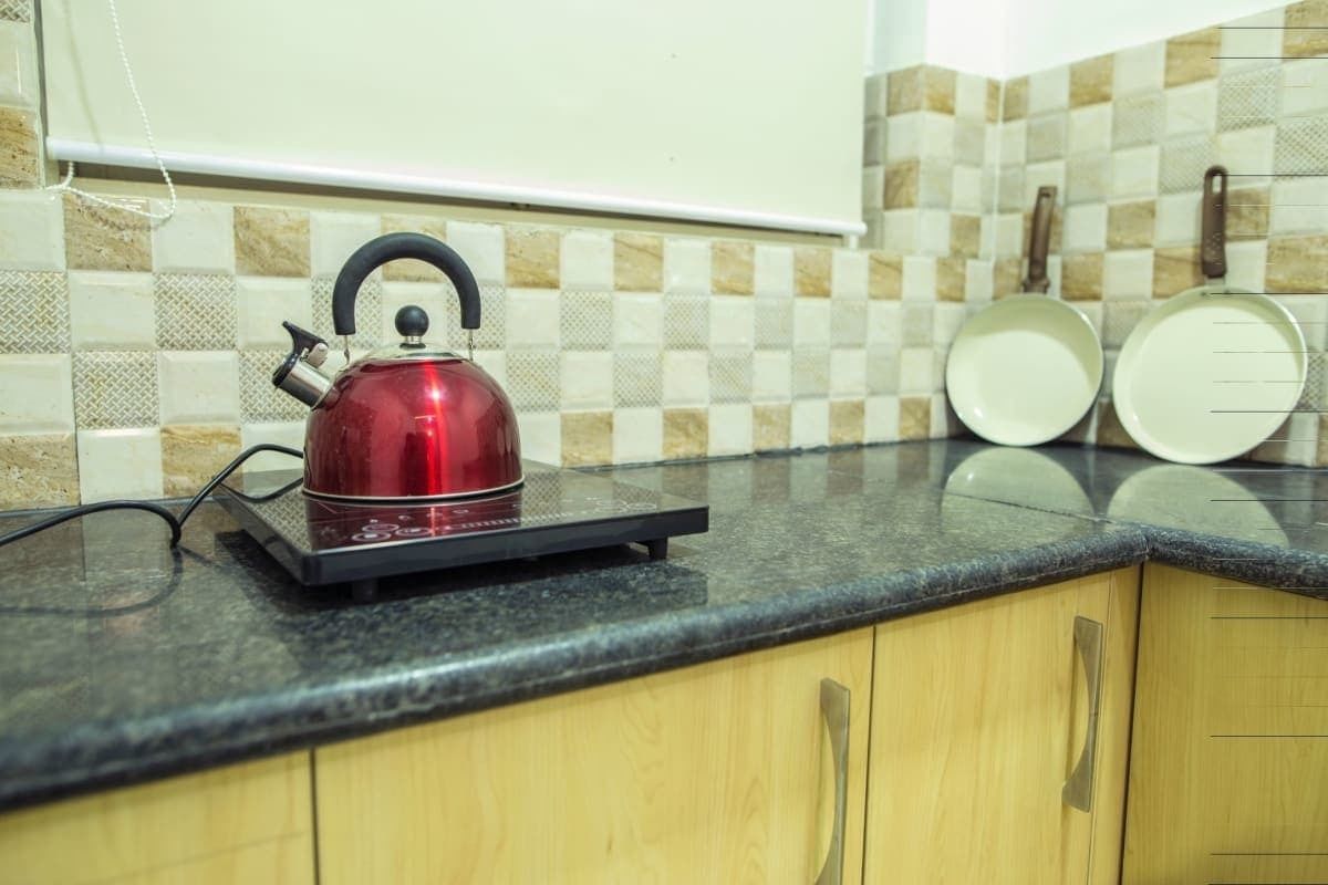 A kitchen with a red kettle on a black induction cooktop, wooden cabinets, and a tiled backsplash.