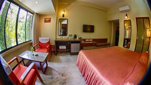 A room with a bed, a desk, a chair, a small table, a television, and a window with curtains. - Hotel Parisutham (Thanjavur)