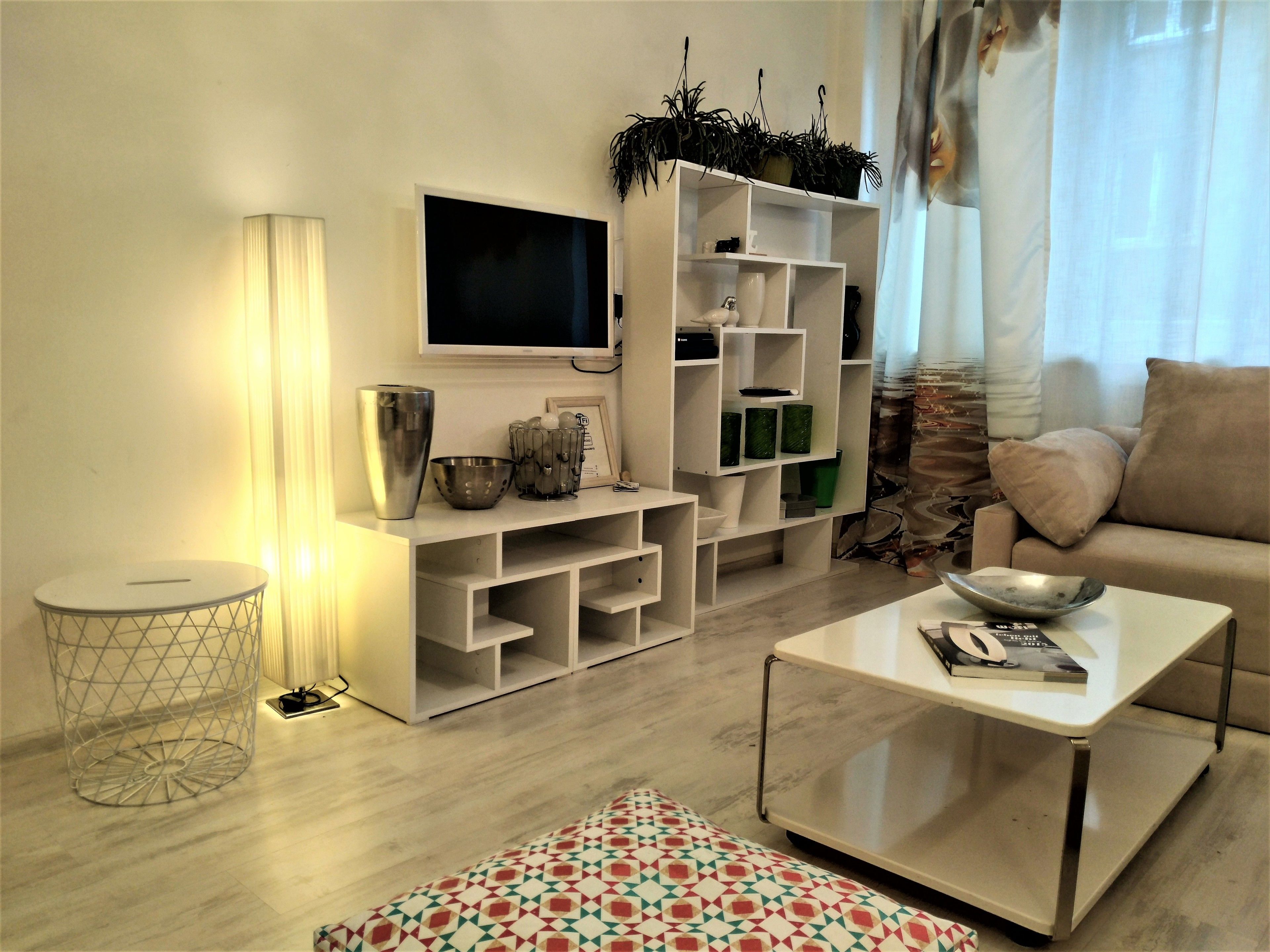 Ein modernes Wohnzimmer mit Fernseher, weißen Regalen, einem Couchtisch und einem gemusterten Hocker.
