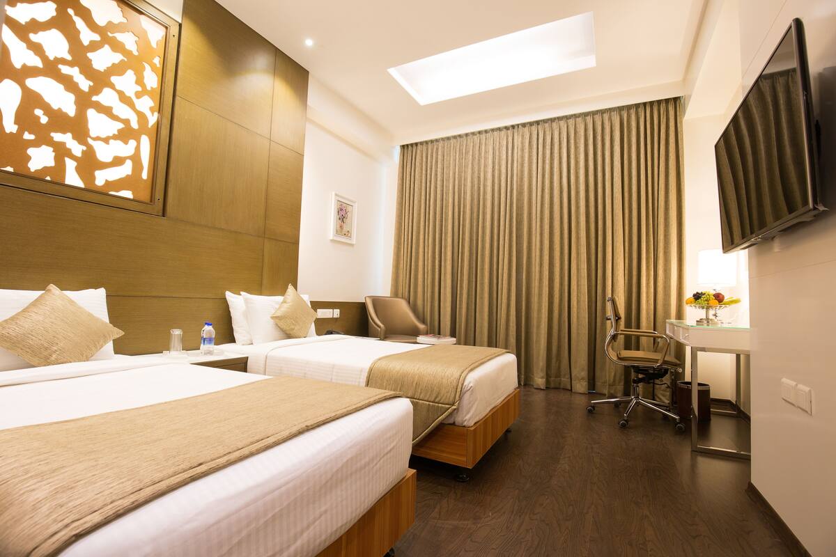 A hotel room with two beds, a desk, a chair, a TV, and a window with curtains.