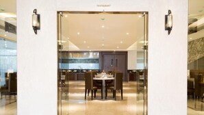 A restaurant with a modern interior, featuring a central dining area with tables and chairs, a kitchen in the background, and a glass entrance. - Hotel Kiscol Grands (Coimbatore)