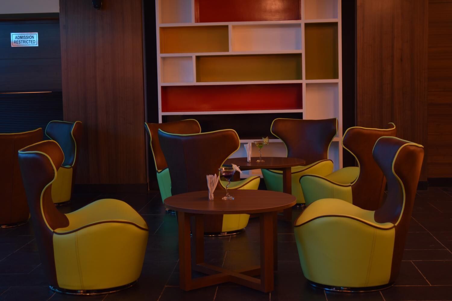 Un'area salotto con sedie colorate e un tavolino, una libreria con volumi di diversi colori, e un cartello che indica l'accesso limitato.