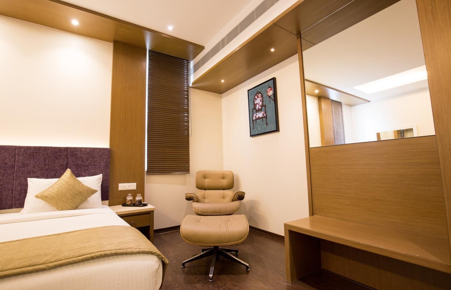 Habitación de hotel con cama, escritorio, silla, mesita de noche, ventana con persianas y un cuadro en la pared.