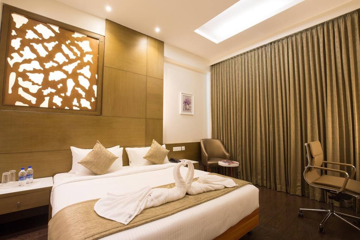 A hotel room with a large bed, a desk with a chair, a small table, and a large window with curtains.
