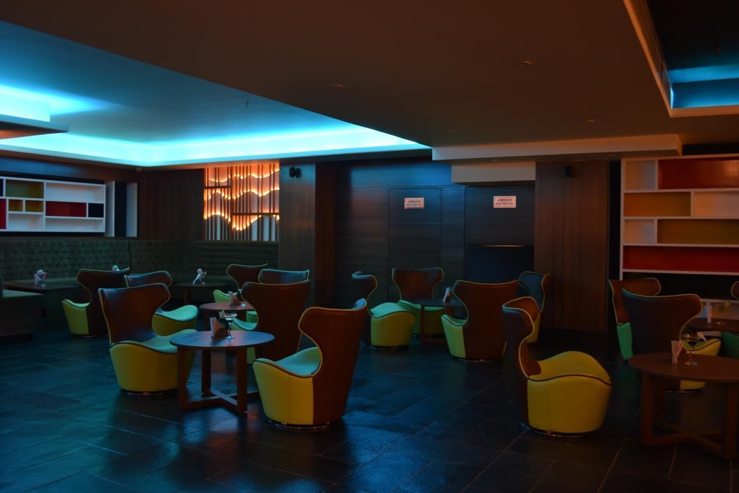 A modern lounge area with colorful chairs and tables, a decorative wall with vertical elements, and a ceiling with blue lighting.