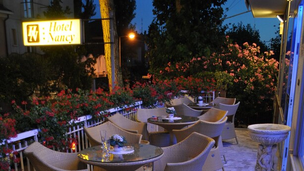 An outdoor patio area with wicker furniture, a stone floor, and a sign for Hotel Nancy. - Hotel Nancy (Rimini)