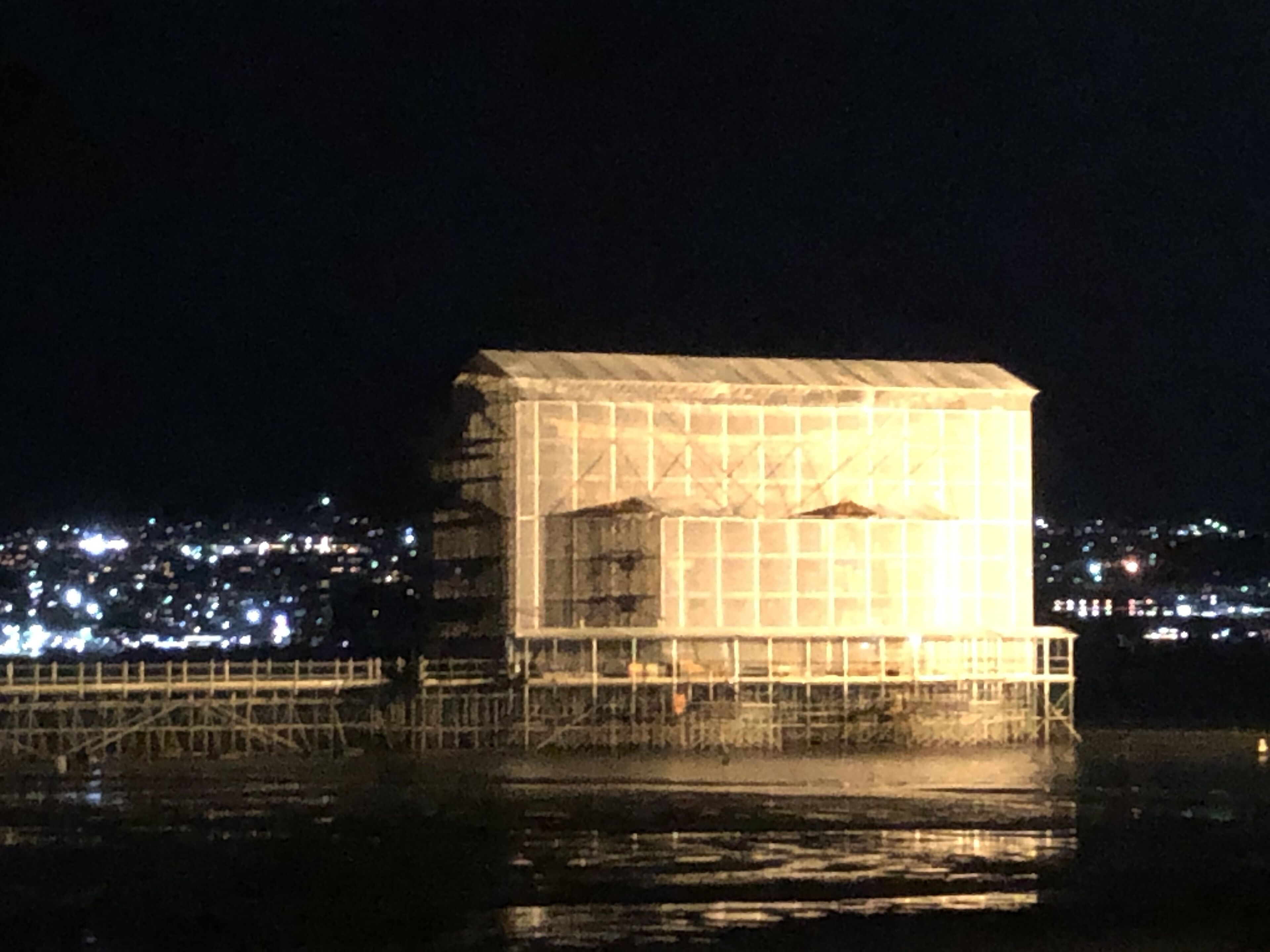 Un edificio grande e iluminado con una fachada de diseño geométrico, ubicado en la ribera de un río con un puente al frente.