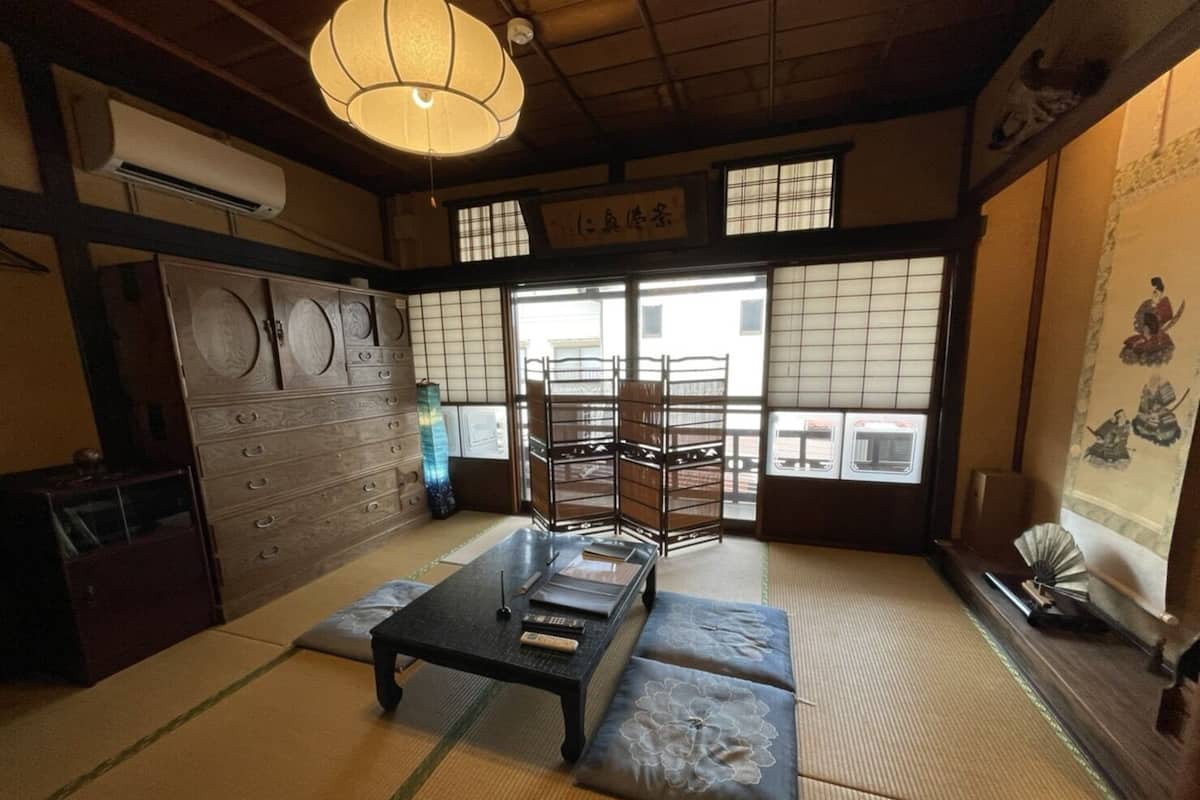 Ein traditioneller japanischer Raum mit Tatami-Boden, einem niedrigen Tisch mit Kissen, einer hängenden Leuchte und einer Schiebetür mit Blick ins Freie.