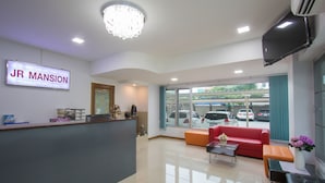 A reception area with a sign reading "JR MANSION," a reception desk, a red sofa, a coffee table, and a window with a view of parked cars. - JR Mansion (Bangkok)