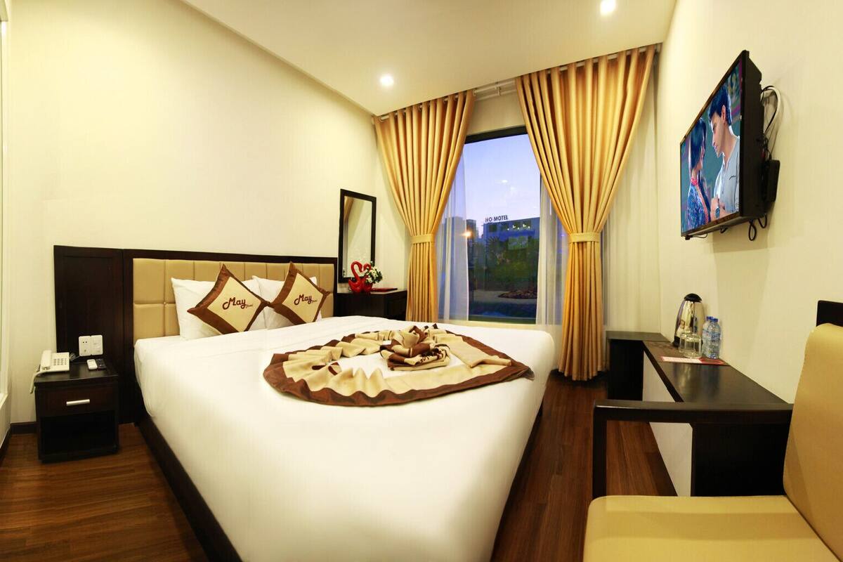 A hotel room with a large bed, a bedside table with a phone, a nightstand, a desk with a chair, a TV on the wall, and a window with curtains.