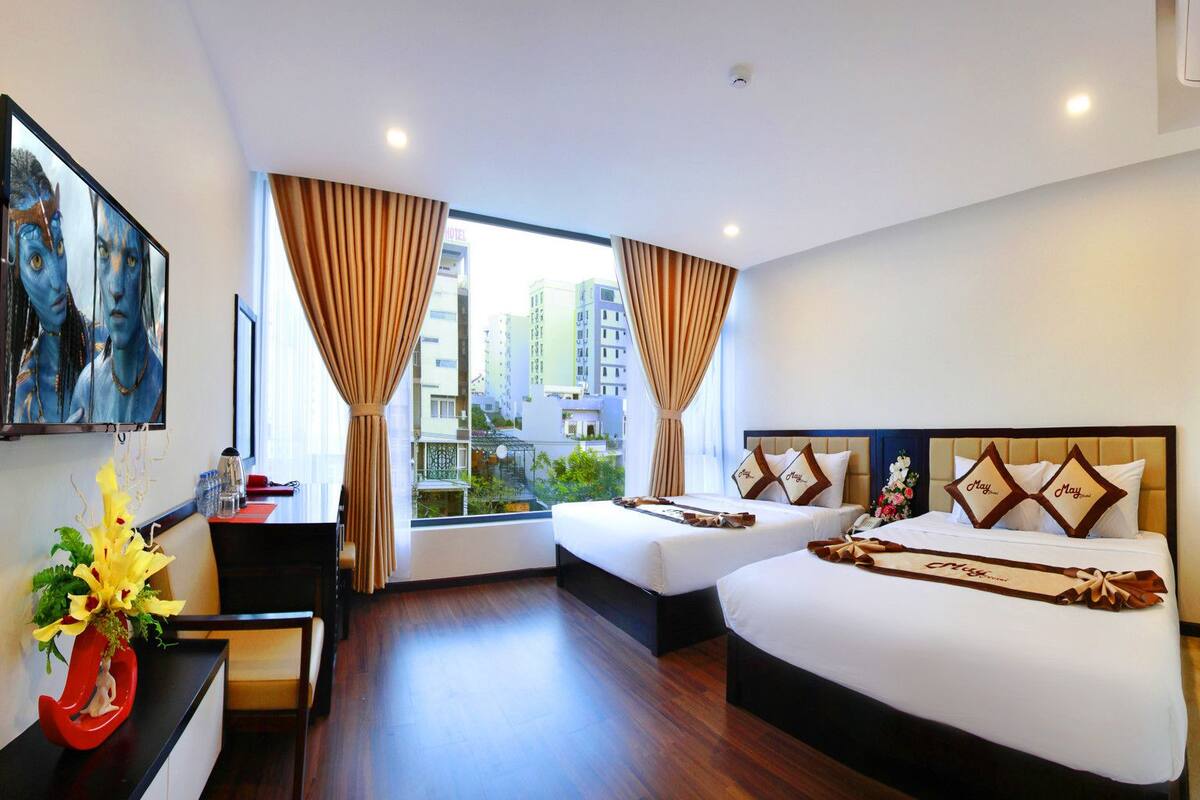 A hotel room with two beds, a desk, a TV, and a large window with curtains.