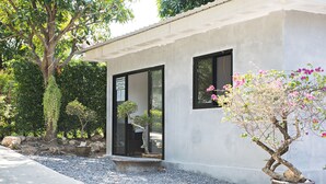 Een gebouw met een betonnen gevel, een deur en een raam. Naast het gebouw staat een pot met een boom met roze bloemen.