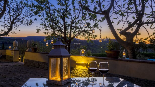A glass table with two wine glasses and a lantern on a patio with potted plants and a view of the city skyline at dusk. - Agriturismo Il Segreto di Pietrafitta (San Gimignano)