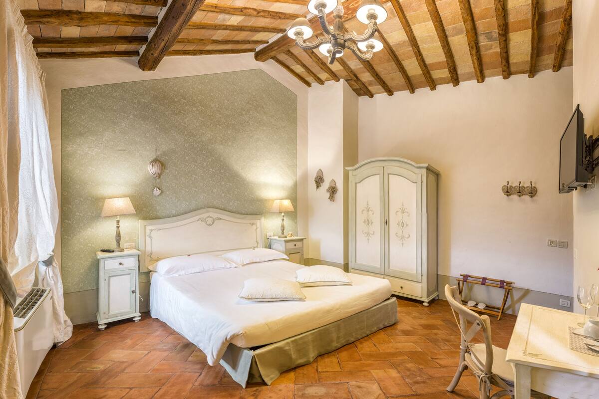 Un dormitorio con una cama grande, dos mesitas de noche con lámparas, un armario, una araña y una mesa de comedor con vasos y un florero.