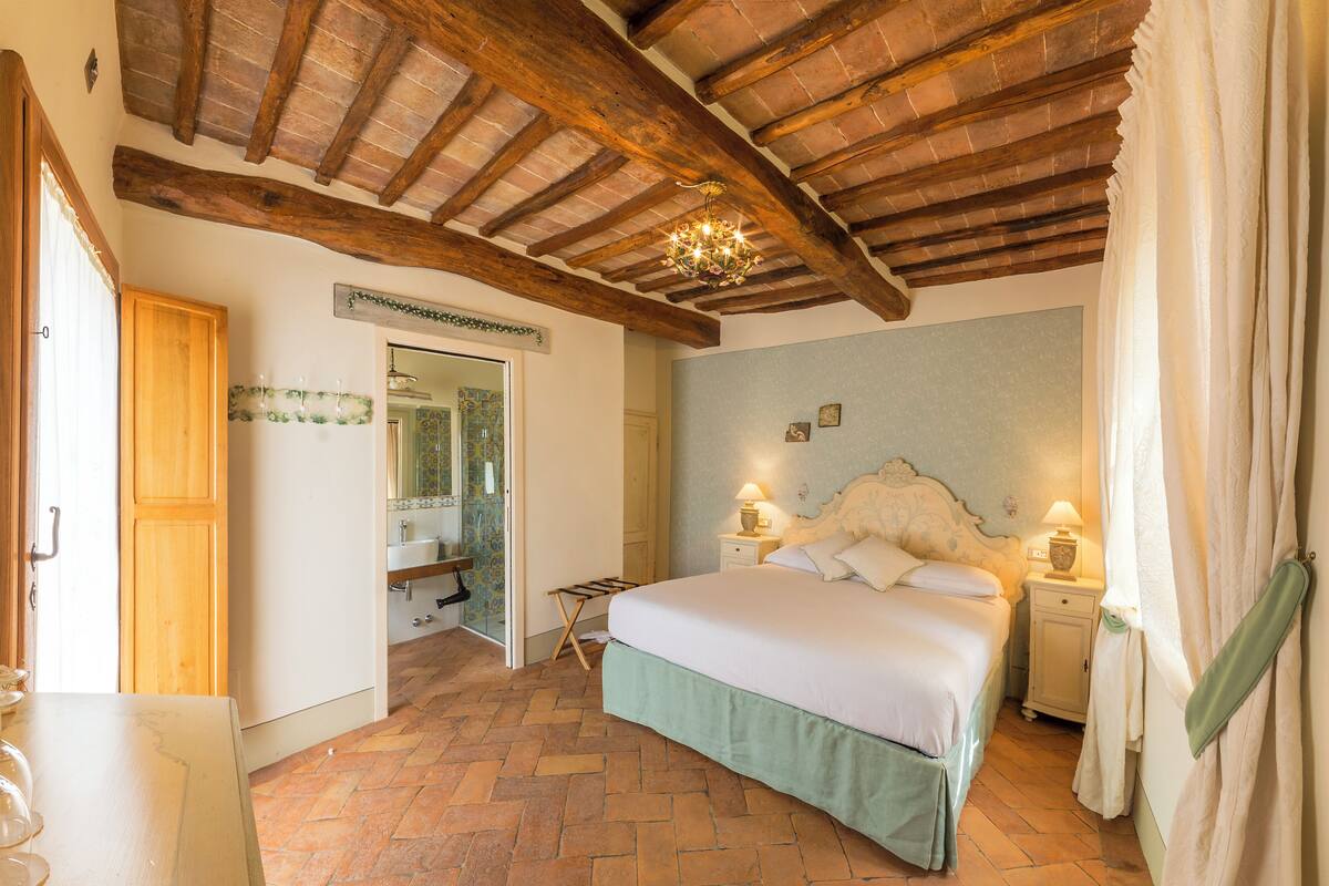 Un dormitorio con techo de madera, una cama con ropa de cama blanca, una mesita de noche con lámpara y una ventana con cortinas.