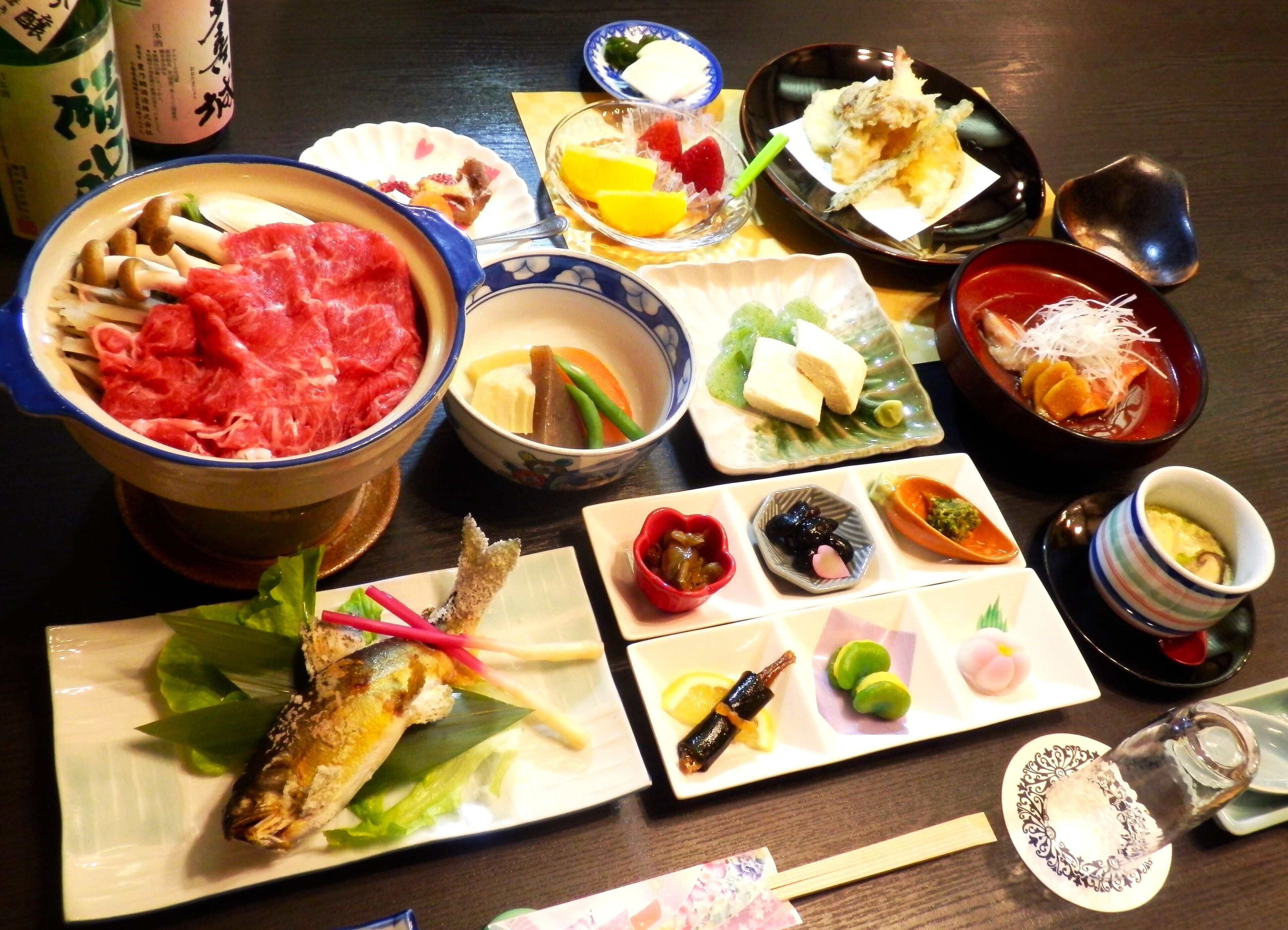 A variety of Japanese dishes including a bowl of raw beef, a fish on a leaf, and several small plates with pickles and vegetables.