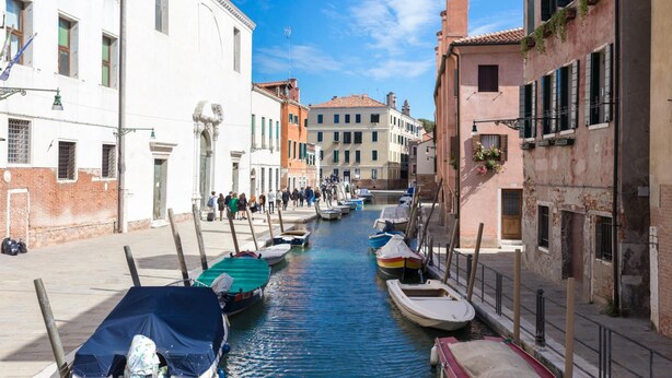 A narrow canal with boats docked on both sides, flanked by historic buildings with balconies and shutters. - InLaguna (Venice)