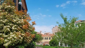 A narrow pathway leading through a lush green area with a brick building on the left and a row of traditional buildings in the background. - InLaguna (Venice)