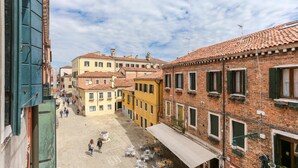 A narrow street with buildings on both sides, featuring a mix of brick and plaster facades, and a small open square with tables and chairs. - InLaguna (Venice)
