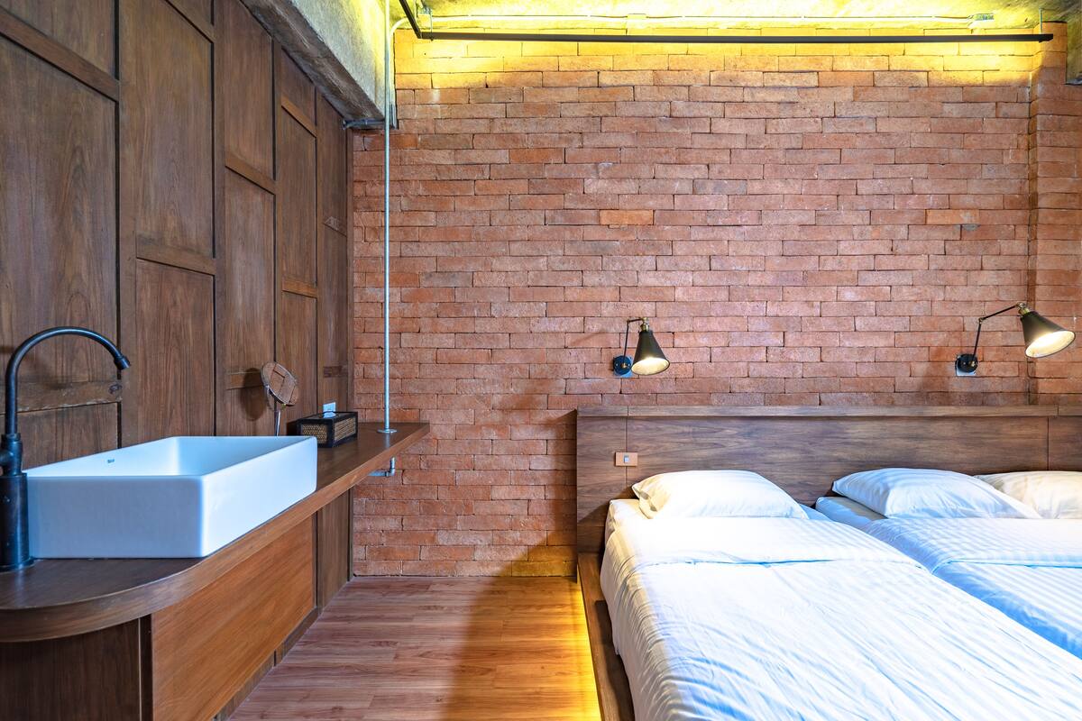 A room with a wooden vanity, a white sink, a brick wall, and a bed with white linens.