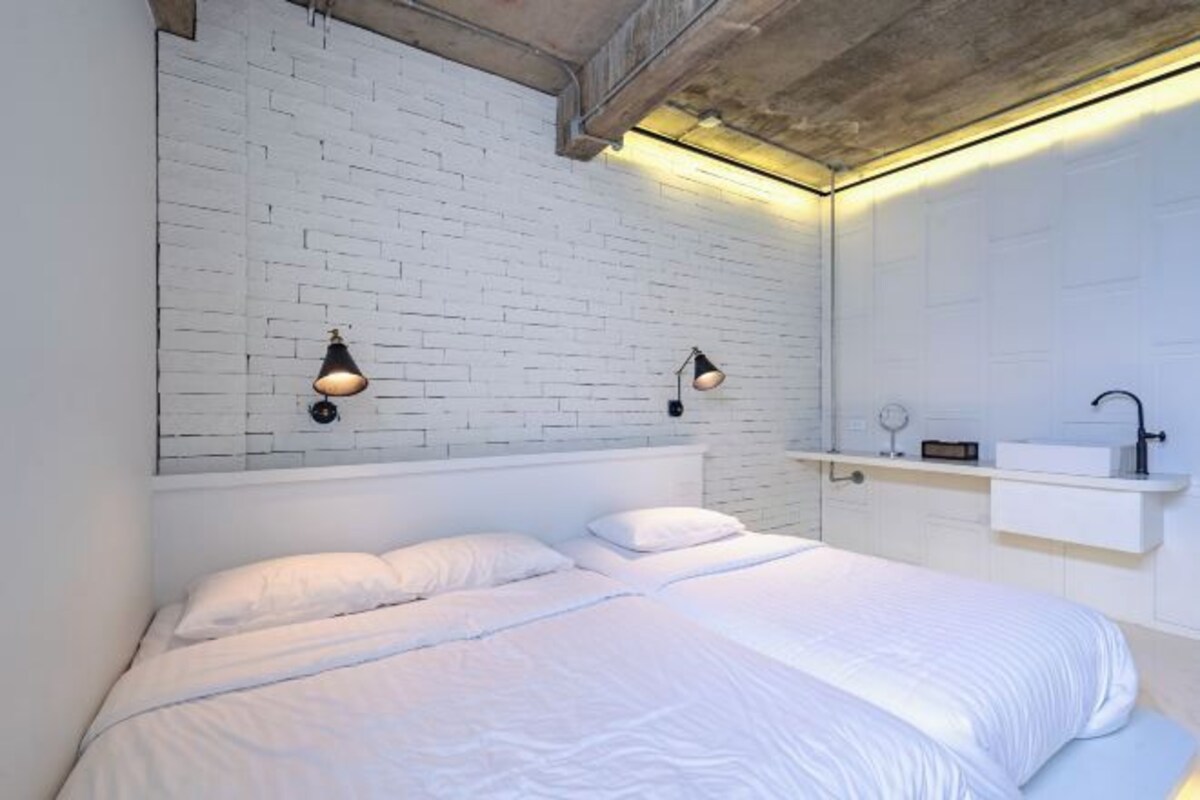 A bedroom with a white brick wall, a bed with white bedding, two wall-mounted lamps, and a white bedside table with a mirror and a radio.