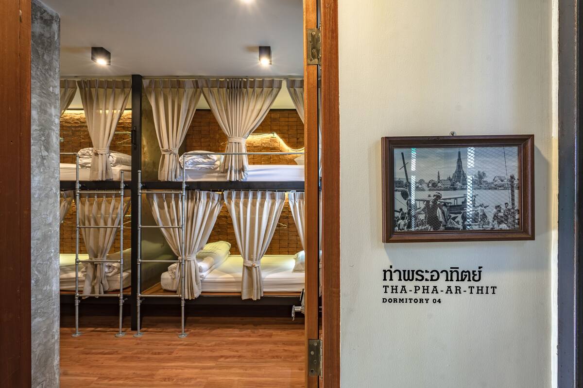 A dormitory room with bunk beds, a framed picture on the wall, and a sign with Thai text.