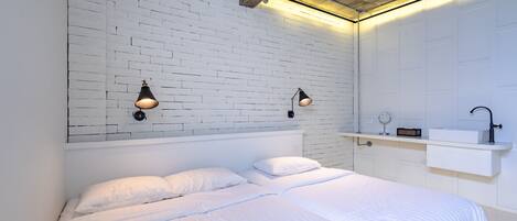 Un dormitorio moderno con pared de ladrillo blanco, dos lámparas de noche, una cama blanca y un pequeño estante blanco con espejo y radio.