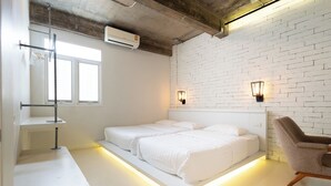 Une chambre d’hôtel moderne avec un mur en briques blanches, deux lits avec du linge de lit blanc, un bureau avec une lampe et une chaise.