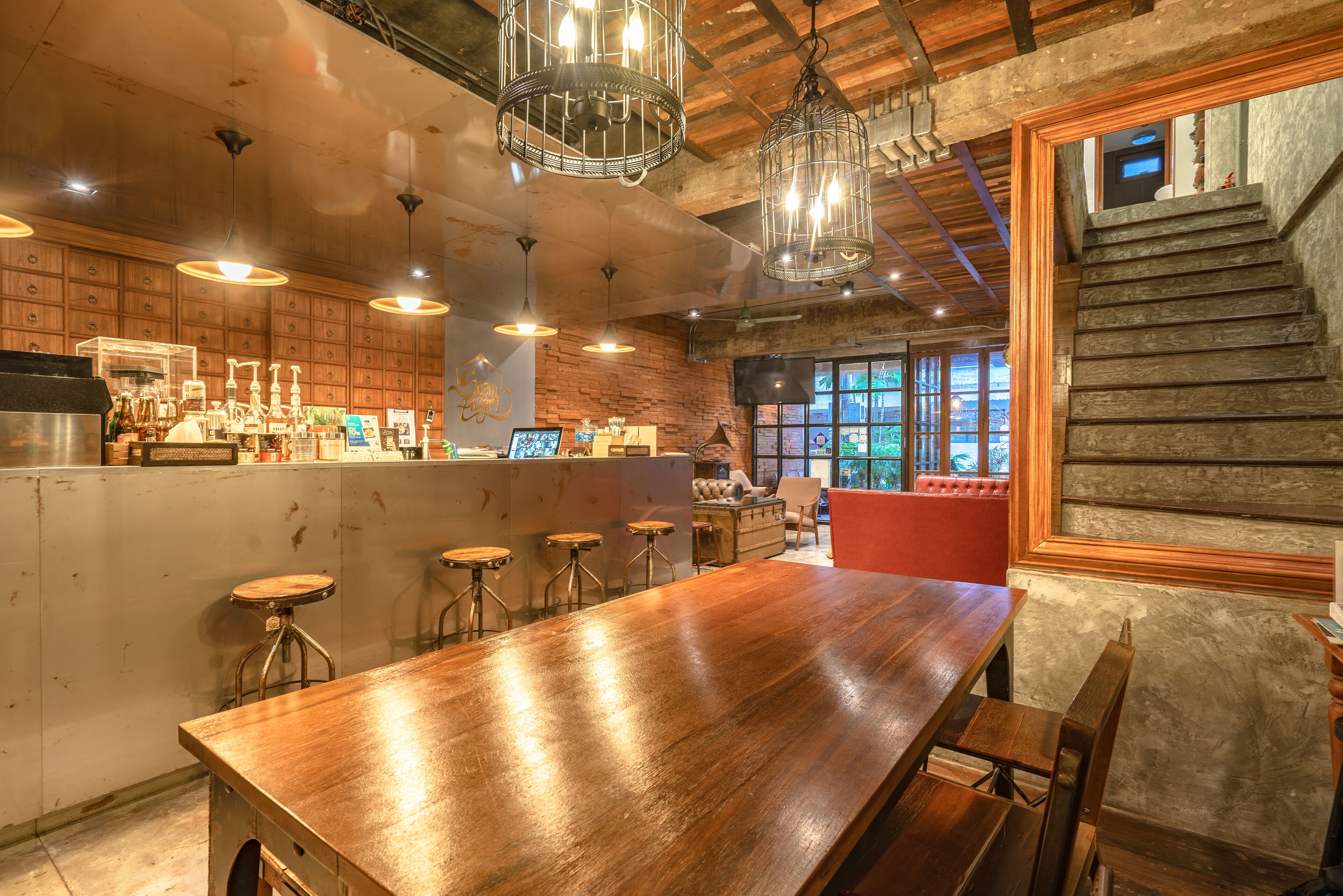 A bar area with a long wooden counter, stools, hanging light fixtures, and a staircase leading to an upper level.