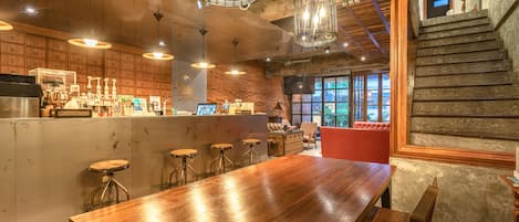 A bar area with a long wooden counter, stools, hanging light fixtures, and a staircase leading to an upper level.