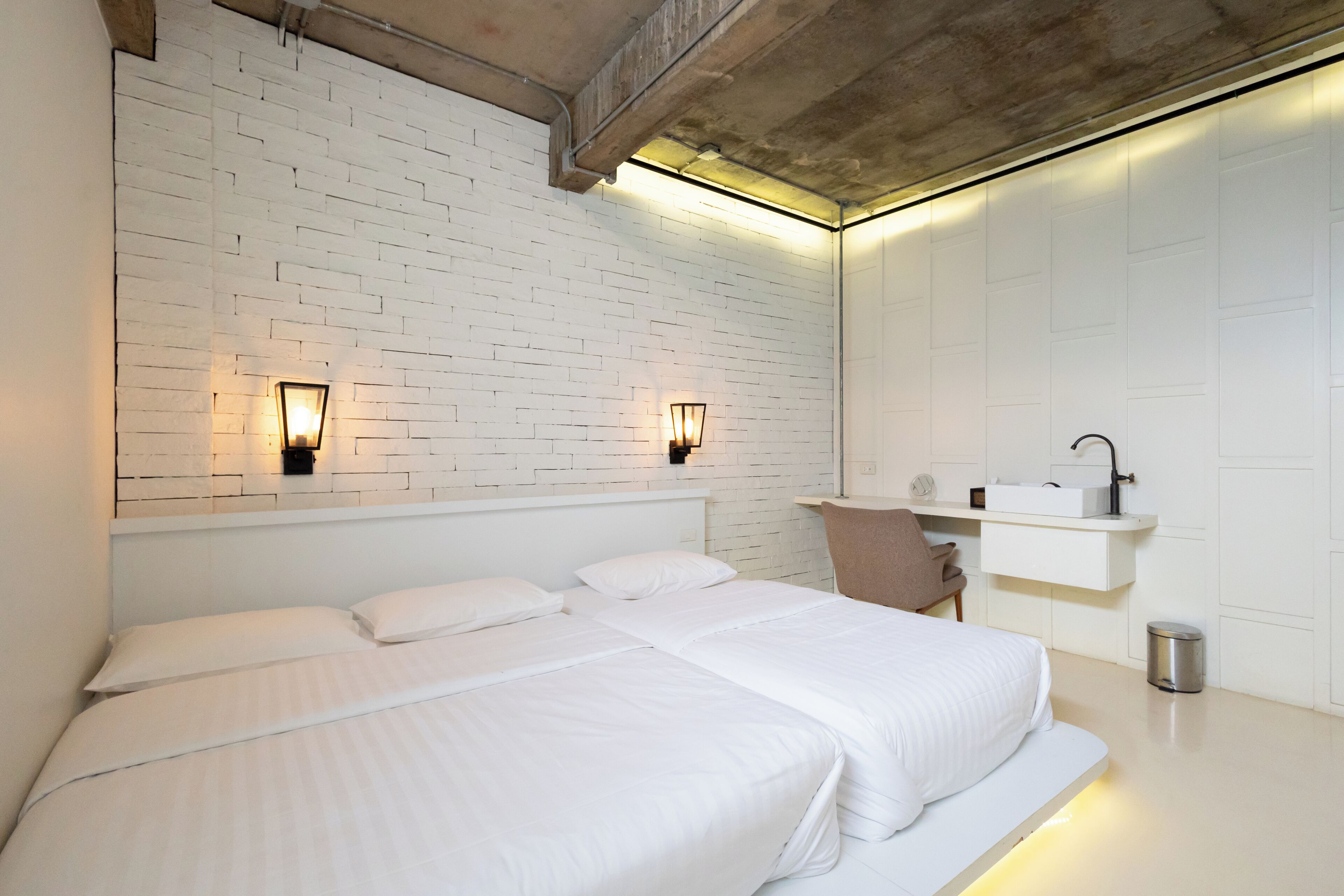 A modern hotel room with a white brick wall, two beds, a small seating area, and a built-in shelf with a mirror and a sink.