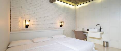 A modern hotel room with a white brick wall, two beds, a small seating area, and a built-in shelf with a mirror and a sink.