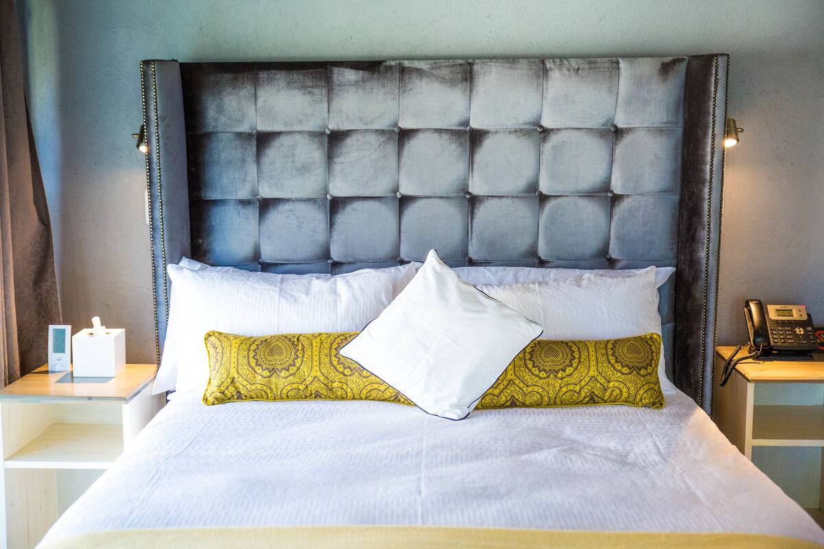 A neatly made bed with a tufted headboard, white and patterned pillows, and a bedside table with a telephone and a tissue box.