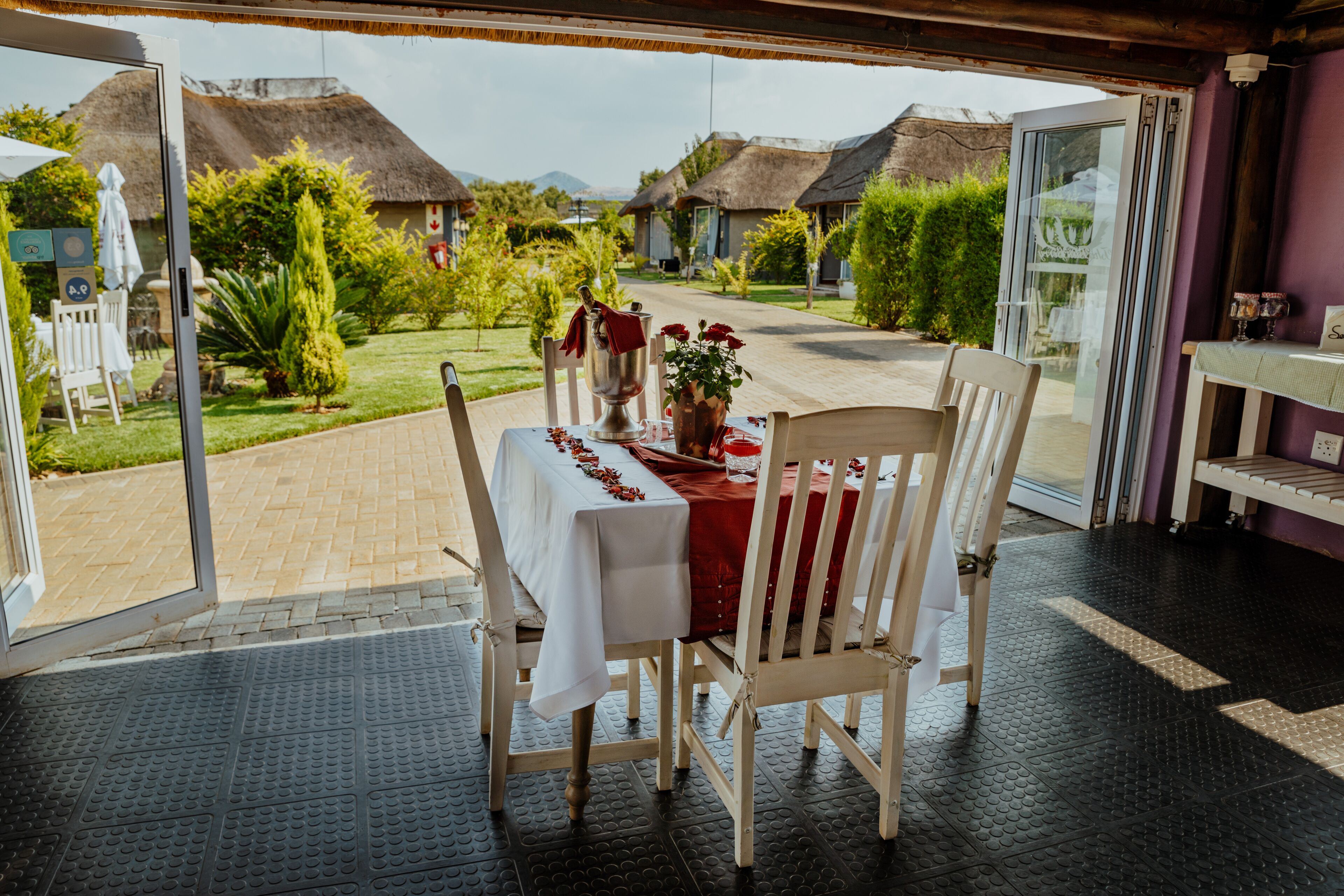 A dining area with a table set for two, overlooking a garden with thatched-roof buildings.