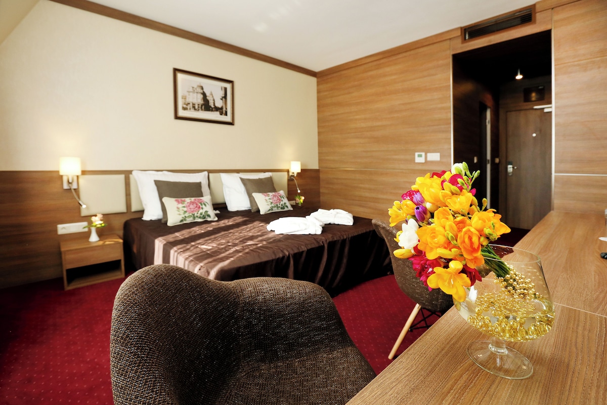 Habitación de hotel con cama, cabecero de madera, mesita de noche con lámpara, una mesa con un florero y un cuadro en la pared.