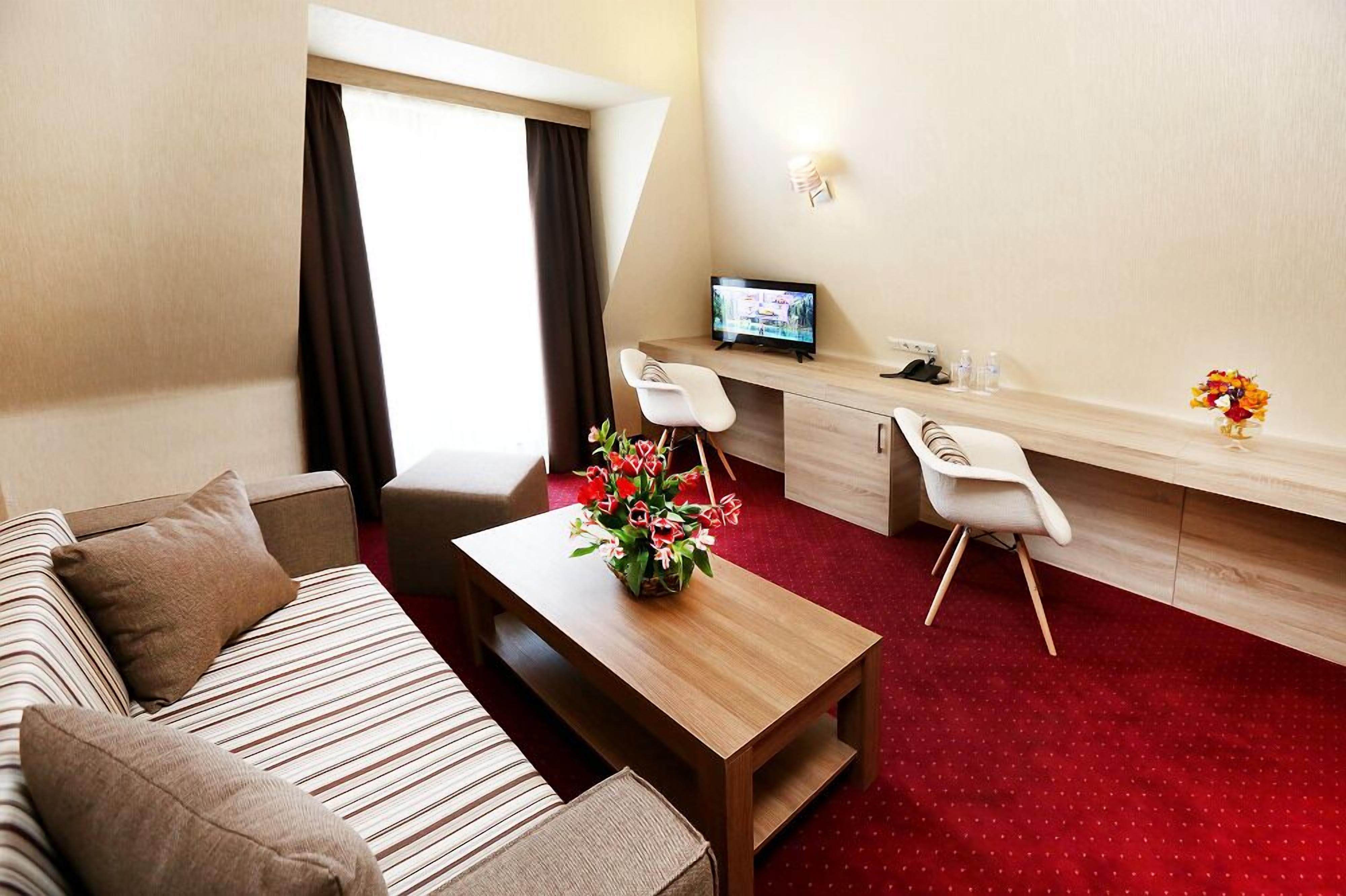 Habitación de hotel con sofá, una mesita de centro de madera con un florero, un escritorio con una computadora y una alfombra roja.
