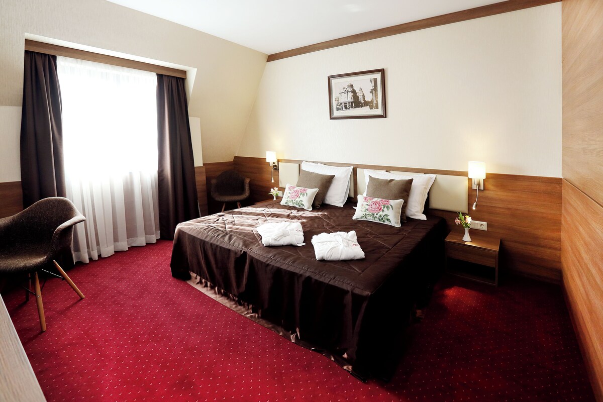 A hotel room with a large bed, two bedside tables with lamps, a chair, a window with curtains, and a framed picture on the wall.