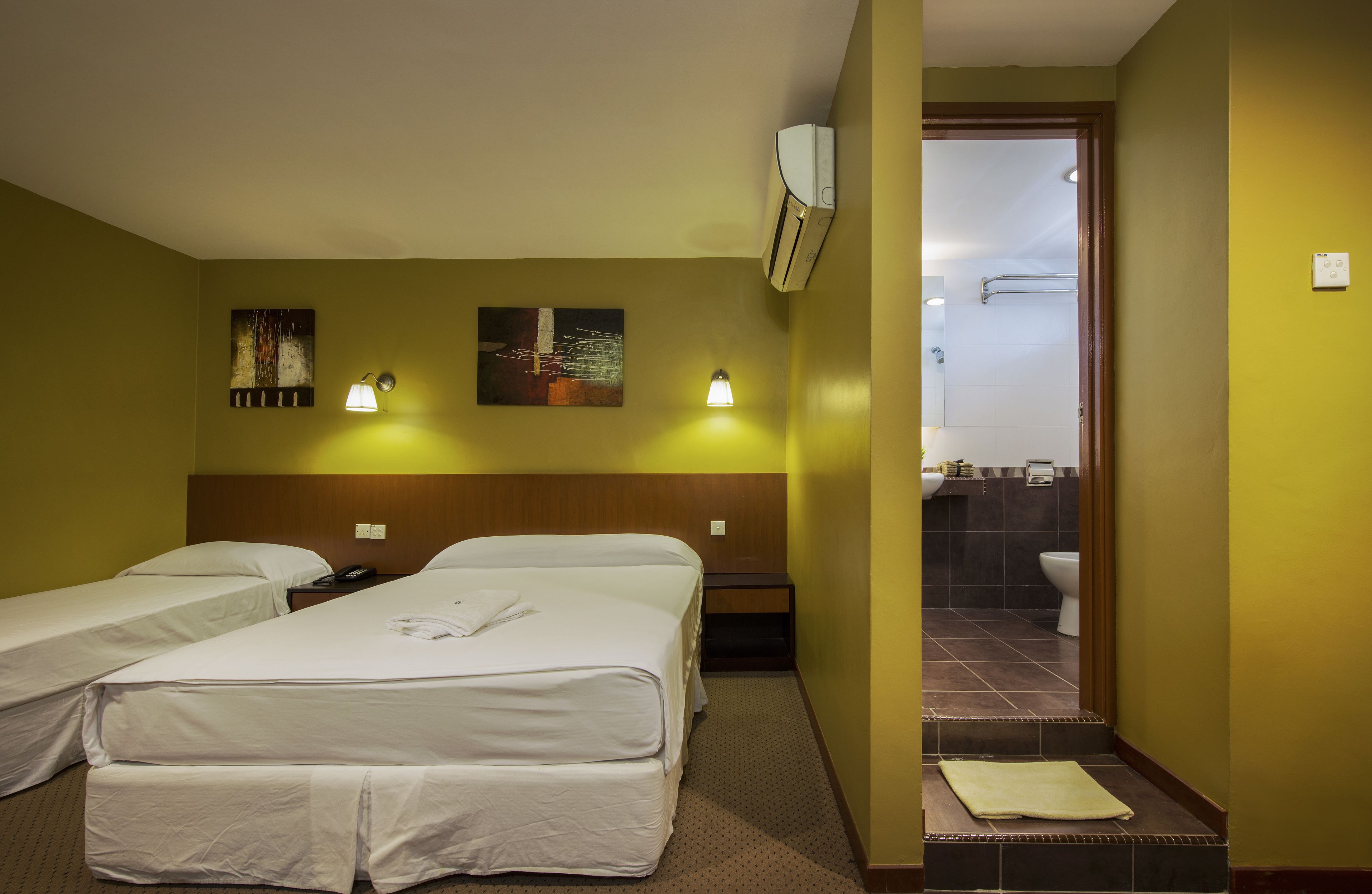 A hotel room with two beds, a nightstand, a wall-mounted air conditioner, and a bathroom visible through an open door.