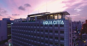 Un hotel llamado AQUA CITTA con un diseño arquitectónico moderno, que presenta una amplia terraza en la azotea y un gran letrero iluminado.