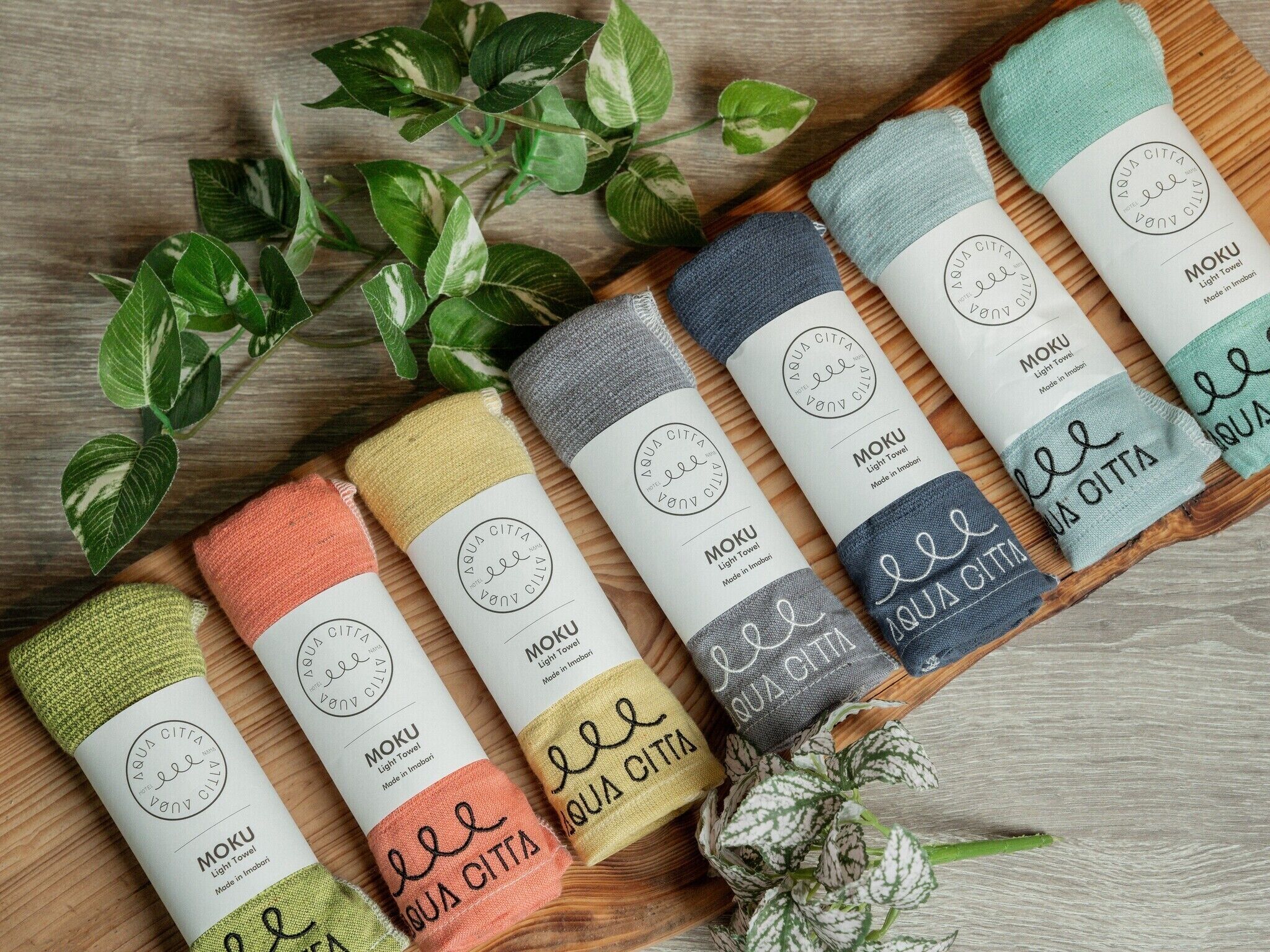 A selection of light-colored towels with the brand name 'MOKU' and 'AQUA CITTA' printed on them, arranged on a wooden surface with green plants in the background.