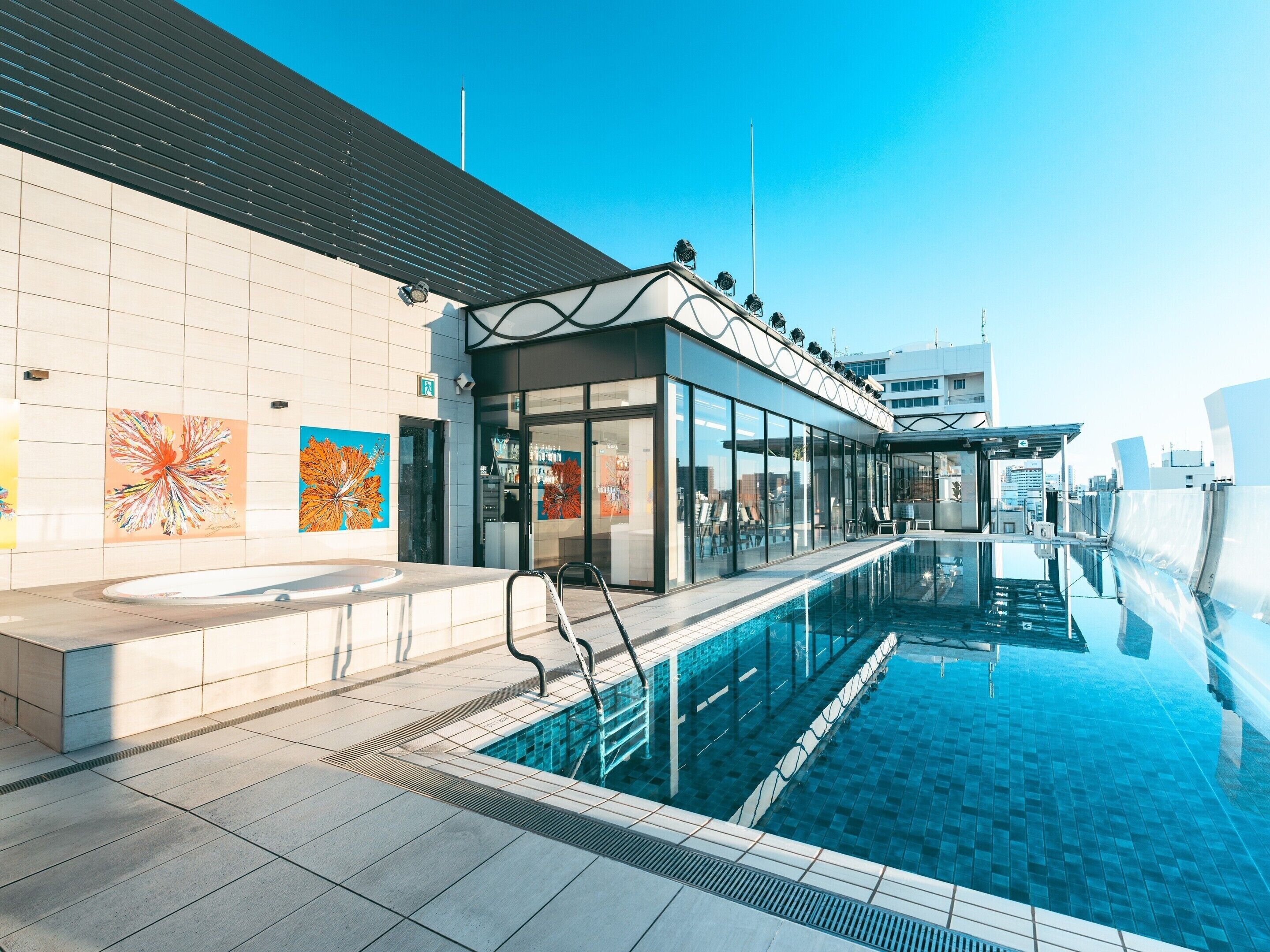 A modern indoor pool area with a clear view of the pool, a lounge area, and a building with large windows and artwork on the wall.