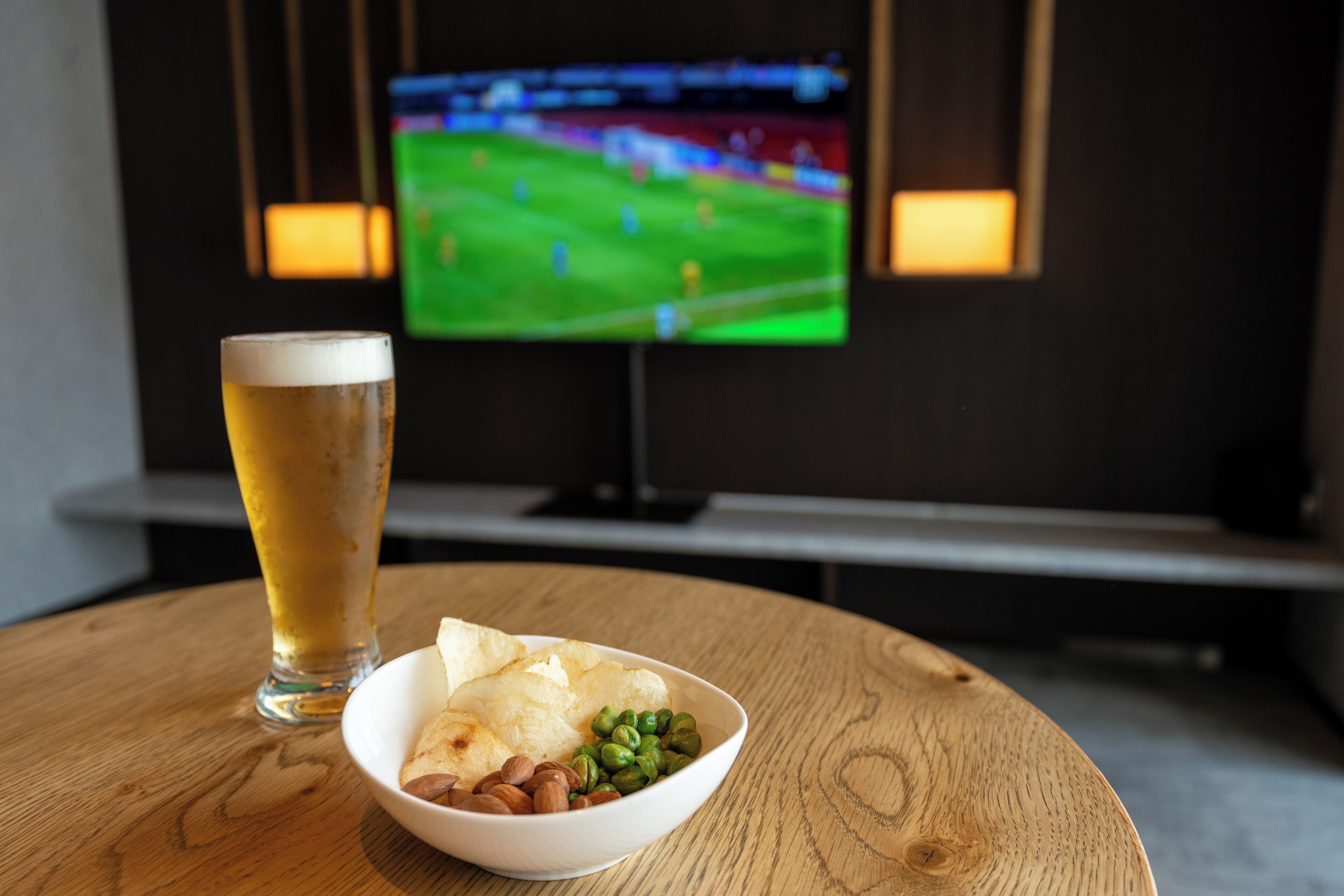 Una mesa con una cerveza en un vaso, un plato de comida y un televisor mostrando un partido de deportes.