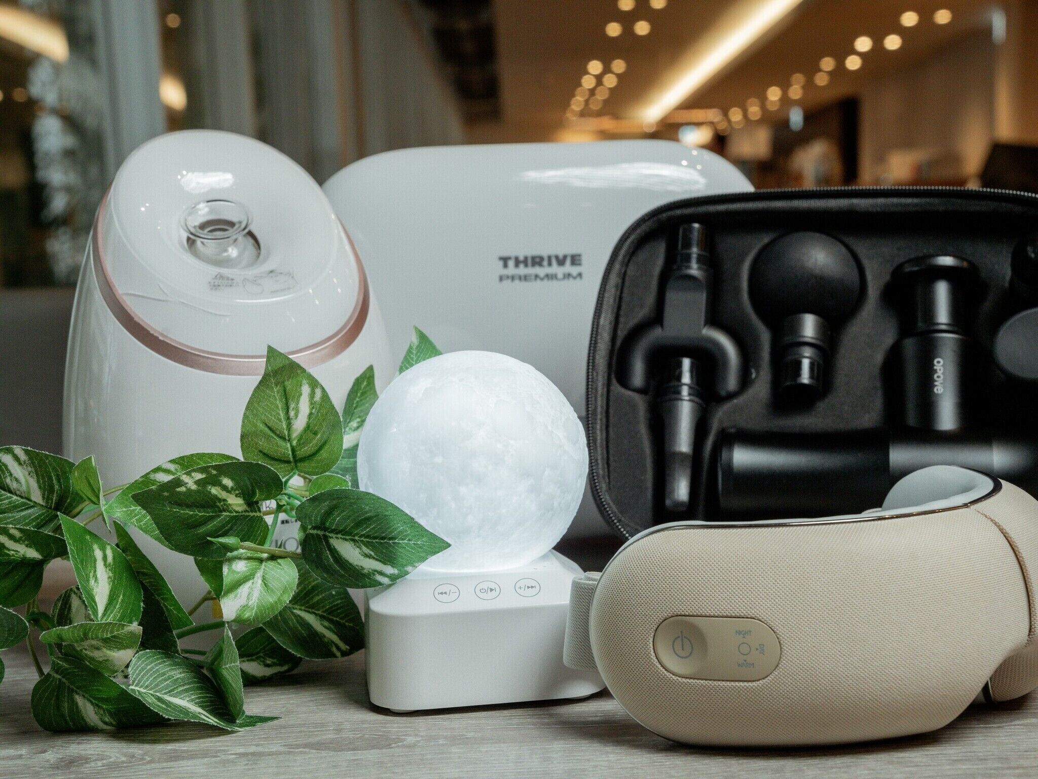 Un dispositivo THRIVE PREMIUM blanco, una funda negra con auriculares y un objeto esférico blanco sobre una superficie de madera con hojas verdes.