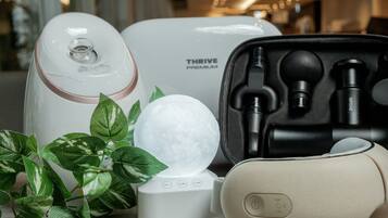 Un dispositivo THRIVE PREMIUM blanco, una funda negra con auriculares y un objeto esférico blanco sobre una superficie de madera con hojas verdes.