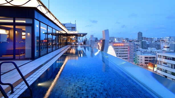 Piscina infinita en la azotea con vistas a la ciudad, barandillas de vidrio y zona de bar.