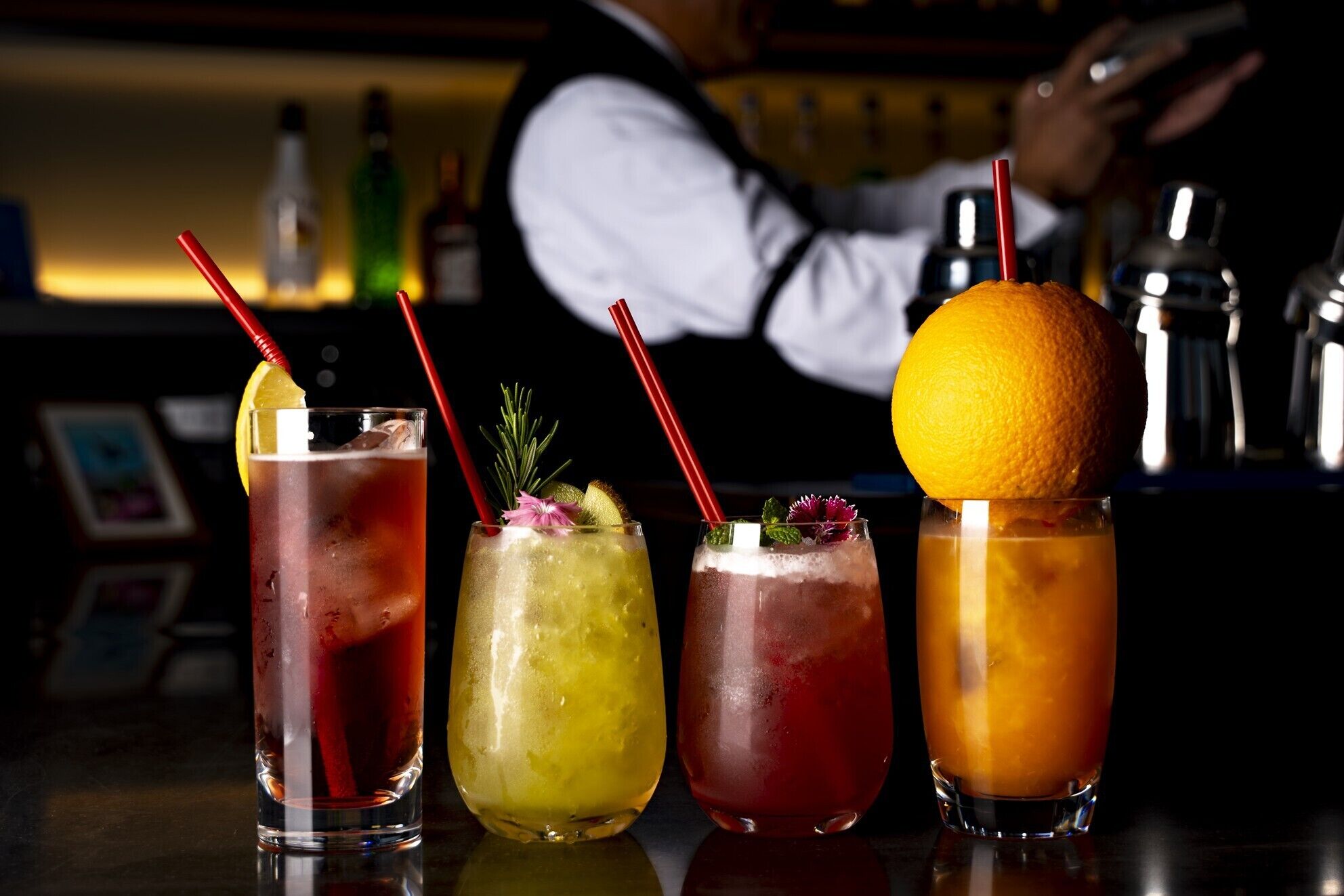 Drei Cocktails mit Garnierungen und Trinkhalmen, im Hintergrund eine Bar und ein Barkeeper.