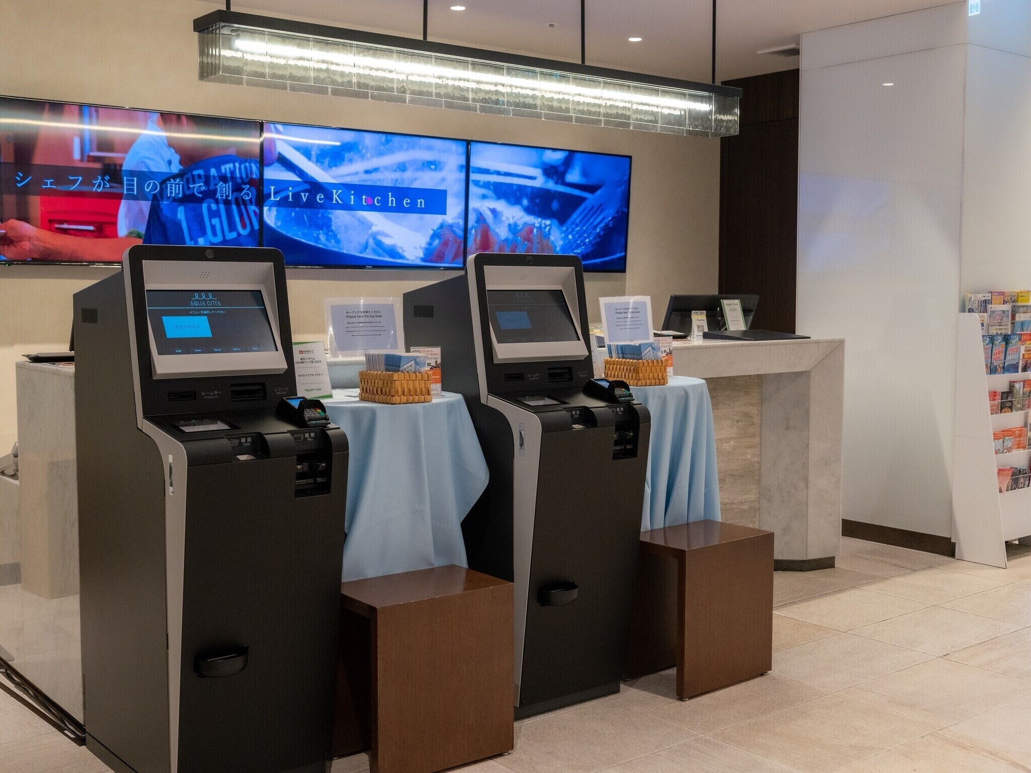 A self-service kiosk area with two kiosks, a digital screen displaying a program, and a counter with informational signs.