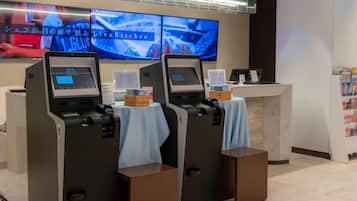 A self-service kiosk area with two kiosks, a digital screen displaying a program, and a counter with informational signs.