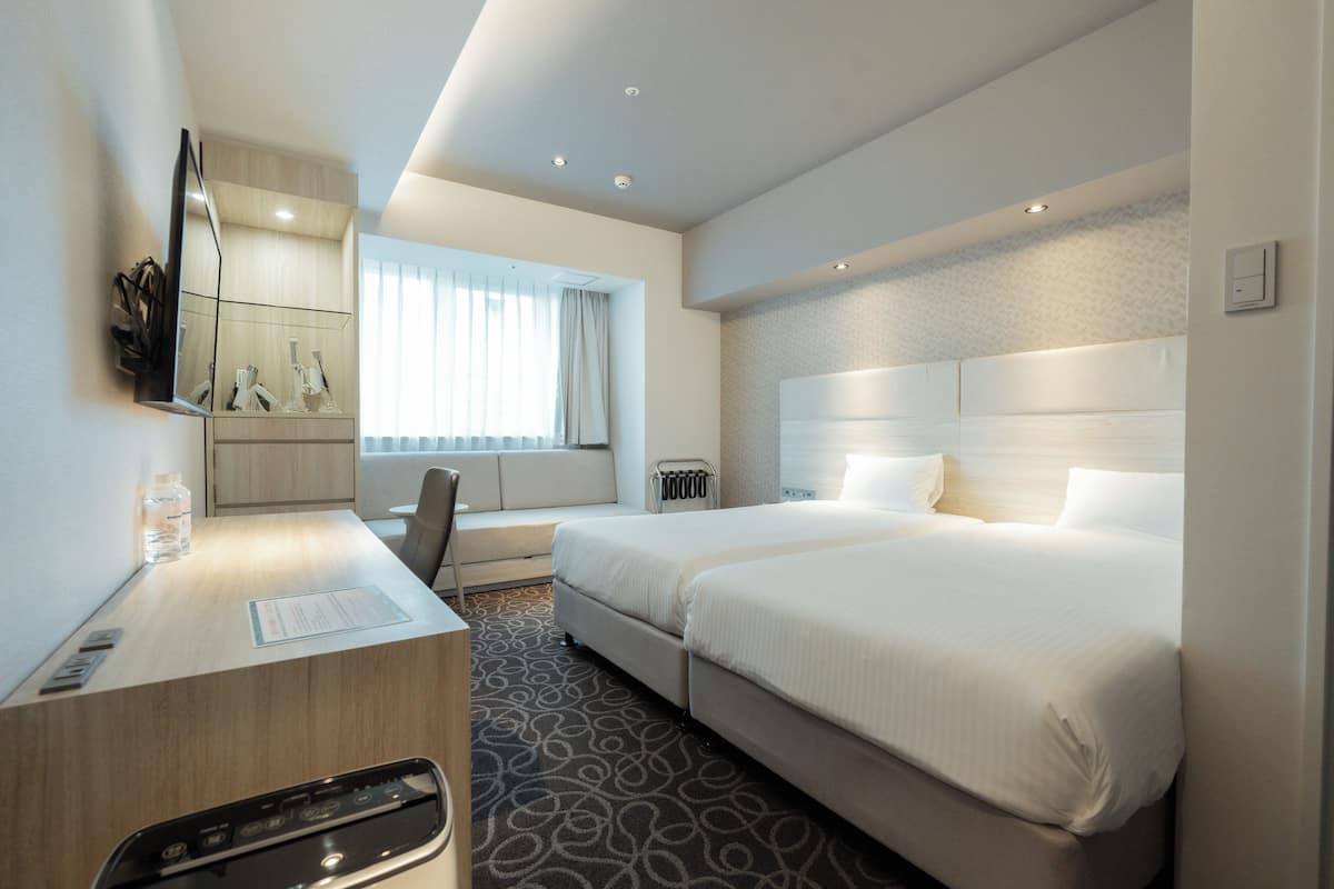 Habitació d'hotel moderna amb escriptori, dos llits, un mirall gran i una finestra amb cortines.